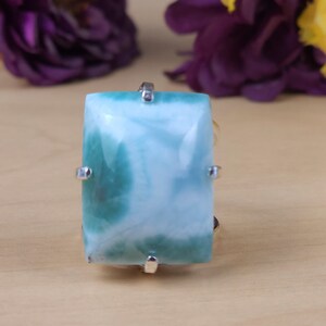 May include: A rectangular larimar gemstone ring featuring a polished, light blue stone with distinct teal and white marbled patterns. The stone is secured by four silver-toned metal prongs, creating a prominent centerpiece for this jewelry item.