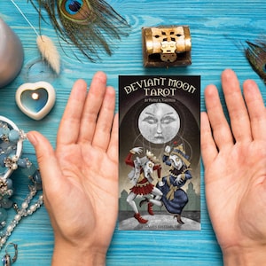 May include: A tarot card from the Deviant Moon Tarot deck, featuring a full moon with a face, and three figures in a dark, whimsical setting. The card is held in the hands of a person.