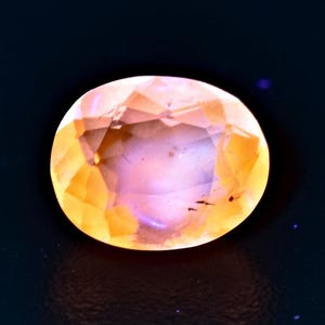 May include: A light pink and orange oval-shaped gemstone with a faceted cut. The gemstone has a translucent appearance and is set against a dark background.