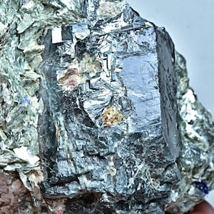 May include: A dark gray, almost black, crystal with a cubic shape, embedded in a matrix of green and gray rock. The crystal has a smooth, reflective surface.