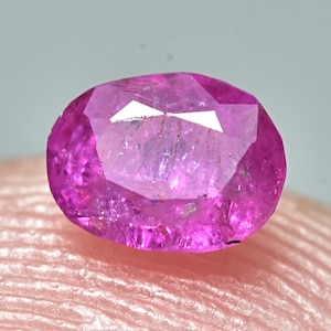 May include: A vibrant pink oval-shaped gemstone with a faceted cut. The gemstone is set against a soft pink background.