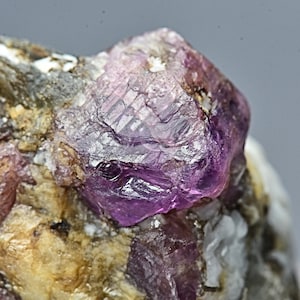 Pode incluir: Um close-up de um cristal de rubi roxo incrustado em uma matriz de rocha cinza e branca. O rubi tem uma forma afiada e facetada e um brilho vítreo.
