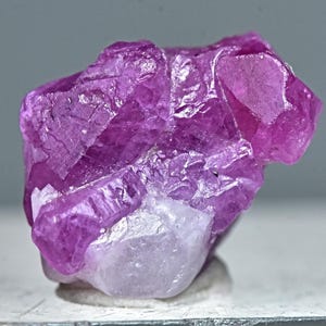 May include: A cluster of pink ruby crystals with a rough, faceted texture. The crystals are translucent and have a deep pink hue.