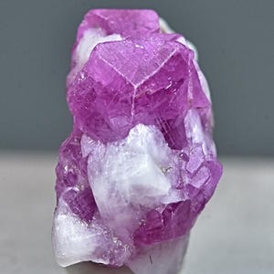Pode incluir: Um aglomerado de cristais de rubi rosa com uma matriz branca. Os cristais são facetados e têm um brilho vítreo.