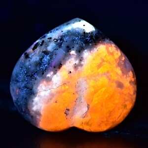 May include: A heart-shaped polished stone with a white and orange pattern. The stone glows under UV light, showing a blue and orange pattern.