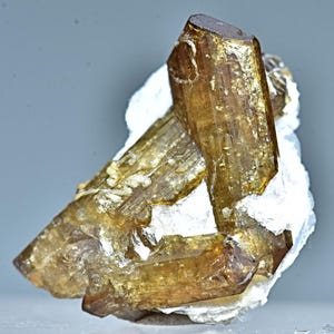 May include: A cluster of brown and green, translucent, elongated crystals with a striated surface. The crystals are arranged in a triangular shape and are set against a white background.