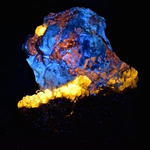 May include: A large, irregularly shaped rock with a blue and orange fluorescent glow under UV light. The rock has a rough, textured surface and is surrounded by smaller, yellow fluorescent crystals.