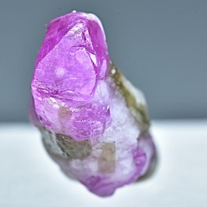 Puede incluir: Un cristal de rubí rosa con un aspecto áspero y natural. El cristal es translúcido y tiene un aspecto ligeramente nublado. El cristal está colocado sobre un fondo blanco.