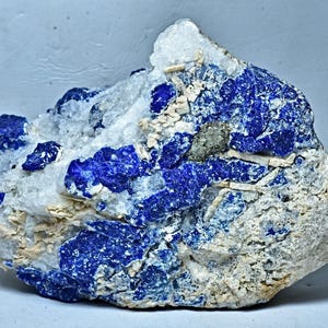 May include: A rough, natural specimen of Lapis Lazuli, a deep blue gemstone, embedded in a white matrix. The stone has a rough, uneven surface and shows a variety of blue hues.