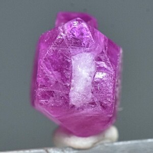 May include: A vibrant pink ruby crystal with a hexagonal shape and a slightly cloudy appearance. The crystal is mounted on a white display stand.