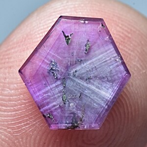 May include: A hexagonal purple sapphire gemstone with a striated surface and inclusions of a darker mineral.