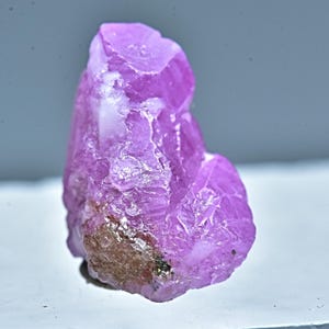 Puede incluir: Un gran cristal de rubí rosa en bruto con un ligero brillo metálico. El cristal tiene una forma natural irregular y es de un color rosa vibrante.