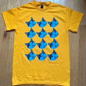 以下が含まれることがあります： マスタードイエローのTシャツ。青い猫の顔が繰り返しプリントされています。猫の顔には黒いディテールがあり、シャツの前面にグリッド状に配置されています。シャツはクルーネックで半袖です。