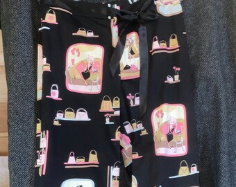 May include: Black pajama pants with a repeating pattern of illustrations featuring women, handbags, and shopping scenes in pink, beige, and white. The pants have a black ribbon tie at the waist.