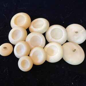 May include: A collection of ten off-white buttons with a raised center and a small hole in the top. The buttons are arranged in a semi-circle on a black surface.