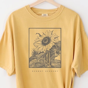 May include: A mustard yellow oversized t-shirt with a black and white illustration of a sunflower. The text "Dermot Kennedy" is printed below the sunflower.