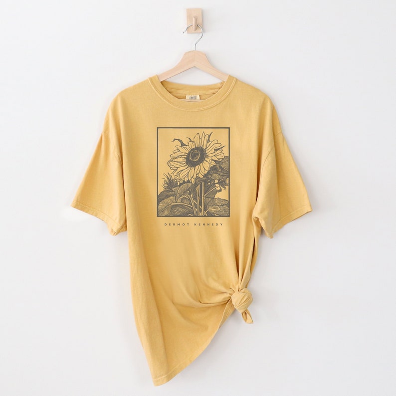 May include: A mustard yellow oversized t-shirt with a black and white illustration of a sunflower. The text "Dermot Kennedy" is printed below the sunflower.