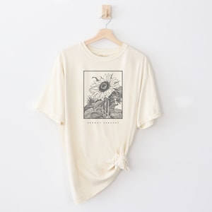 May include: A cream colored t-shirt with a black and white illustration of a sunflower. The text "Dermot Kennedy" is printed below the sunflower.