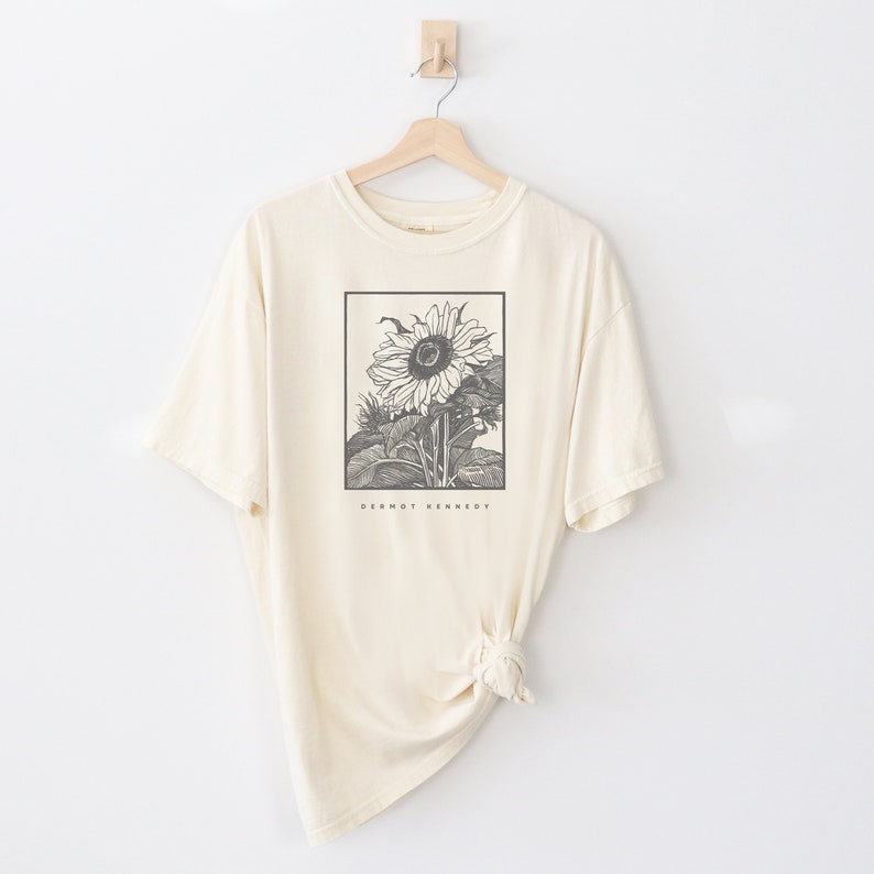 May include: A cream colored t-shirt with a black and white illustration of a sunflower. The text "Dermot Kennedy" is printed below the sunflower.
