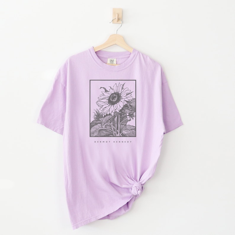 May include: A lavender t-shirt with a black and white illustration of a sunflower in a frame. The text "Dermot Kennedy" is printed below the image.