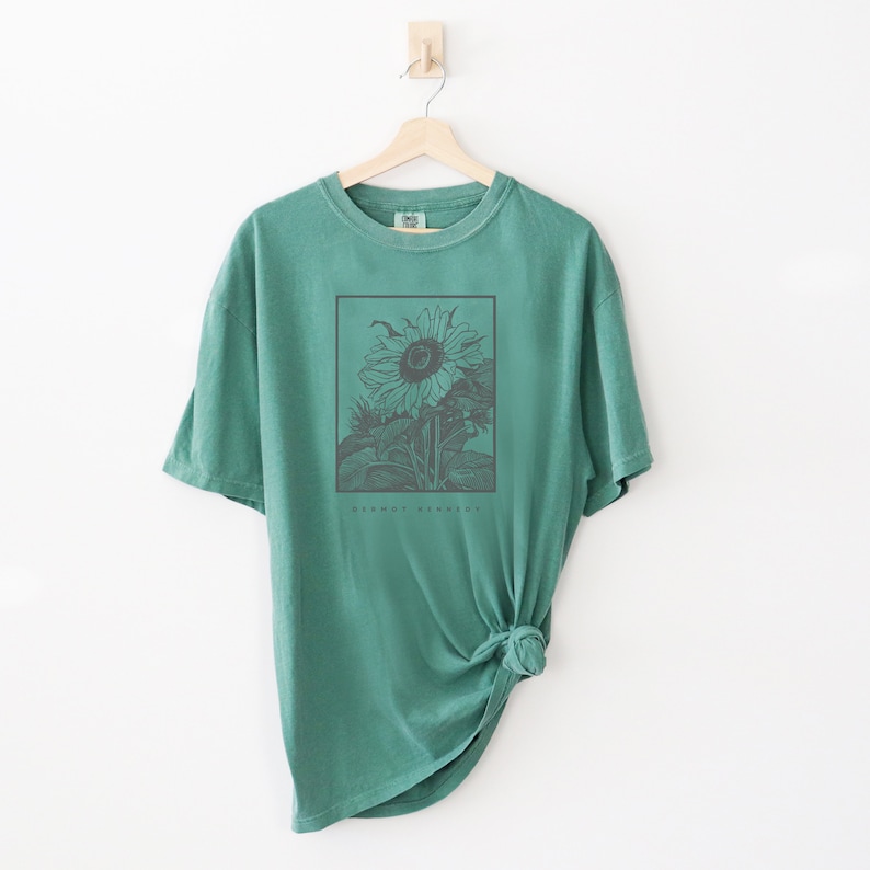 May include: A teal green t-shirt with a black and white illustration of a sunflower. The text "GERNOT RENARD" is printed below the sunflower.