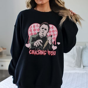 May include: Black sweatshirt with a graphic of a horror movie character holding a knife inside a pink heart with the text "Chasing You".
