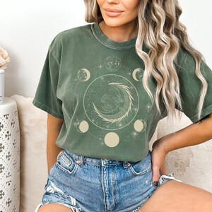 May include: A green t-shirt with a graphic of a crescent moon and stars in a circle. The moon is surrounded by leaves and the graphic is in a beige color.