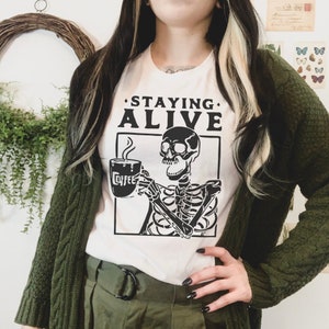 May include: A white short-sleeved t-shirt featuring a black graphic of a skeleton holding a steaming mug labeled COFFEE. Above the skeleton, the text STAYING ALIVE is printed in bold black capital letters. The shirt is worn under a dark green cardigan.