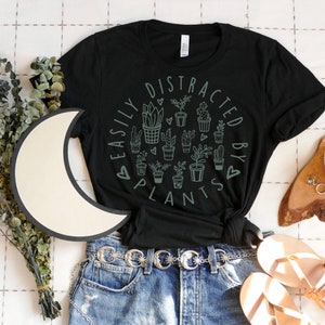 May include: A black short-sleeve t-shirt featuring a circular graphic of various potted plants and cacti in sage green. The text "EASILY DISTRACTED BY PLANTS" is printed in a circular pattern around the plants, accented with small heart symbols.