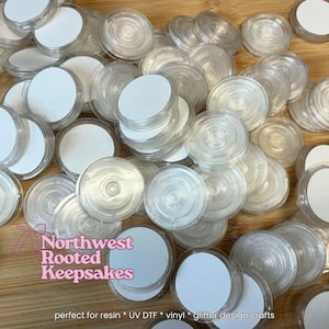 May include: A collection of clear, circular resin craft containers with white circular inserts. The containers are stacked on a wooden surface. The text "Northwest Rooted Keepsakes" is visible in the image.