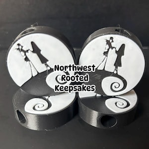 Op de afbeelding: Vier zwart-witte ornamenten met een silhouet van een stel op een heuvel onder een volle maan. De ornamenten hebben een cirkelvormig ontwerp met een spiraalvormige basis en de tekst "Northwest Rooted Keepsakes".
