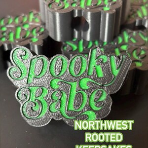 Op de afbeelding: Een zwart en groen "Spooky Babe" aandenken. De tekst is in een gestileerd lettertype met een groene omlijning en vulling. Het aandenken heeft de vorm van een bloem. De tekst "Northwest Rooted Keepsakes" staat eronder.