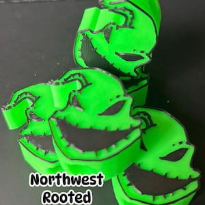 Op de afbeelding: Felgroene, 3D-geprinte Oogie Boogie-thema aandenkens met zwarte accenten. De aandenkens hebben een lachend gezicht met gestikte details. De tekst "Northwest Rooted Keepsakes" is zichtbaar in de afbeelding.