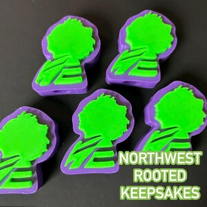 Op de afbeelding: Vijf paarse en groene boomvormige aandenkens. Het boomontwerp is groen, met een paarse omtrek en basis. De tekst "NORTHWEST ROOTED KEEPSAKES" wordt weergegeven in groen en geel.