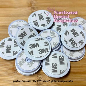 May include: A collection of white round adhesive phone grips with a 3M adhesive backing. The grips are arranged on a wooden surface. The text "3M" is visible on the adhesive backing. The text "Northwest Rooted Keepsakes" is also visible.