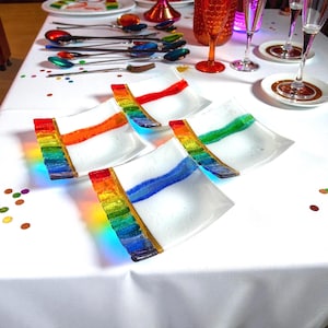 Puede incluir: Platos cuadrados de vidrio blanco con diseños coloridos de arcoíris y rayas. Los platos están colocados sobre un mantel blanco, junto con cubiertos y cristalería coloridos, lo que sugiere un ambiente festivo.