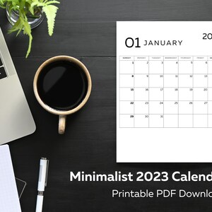 May include: A minimalist calendar for January 2023, with a black and white design. The calendar is on a dark wood surface with a cup of coffee, a laptop, and a plant.