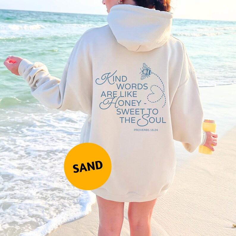 May include: A cream-coloured hoodie with a blue graphic that says "Kind words are like honey - sweet to the soul" and "Proverbs 16:24". The hoodie is being worn by a person standing on a sandy beach.
