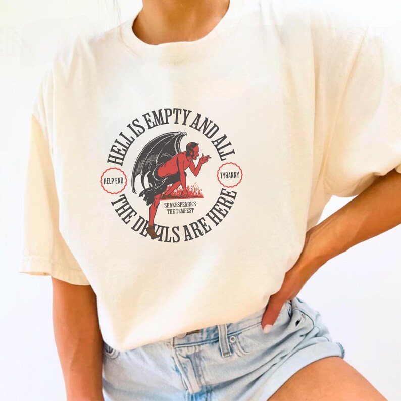 Puede incluir: Camiseta color crema con un dise&ntilde;o de diablo rojo con alas negras y el texto "HELL IS EMPTY AND ALL THE DEVILS ARE HERE." El dise&ntilde;o incluye las palabras "HELP END" y "TYRANNY" en c&iacute;rculos rojos. Una prenda casual.