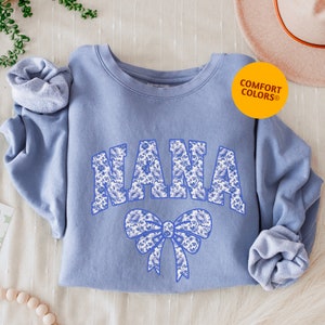 May include: A light blue sweatshirt with the word "NANA" in a floral design across the chest. A matching floral bow is below the word. The sweatshirt has rolled-up sleeves and a "Comfort Colors" logo.