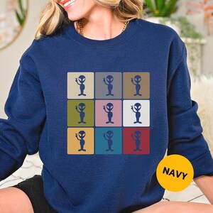 May include: Navy blue sweatshirt featuring a grid of nine colorful squares, each with a cartoon alien illustration. The aliens are in various colors, with a peace sign. A yellow circle with the word "NAVY" is in the bottom right corner.