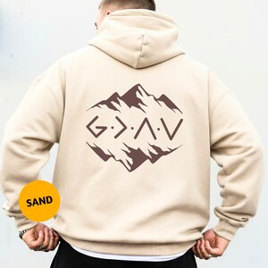 May include: Sand-colored hoodie with a mountain graphic and the letters G > A V in brown. The hoodie has a hood and a kangaroo pocket. The word "SAND" is in a yellow circle in the lower left corner.