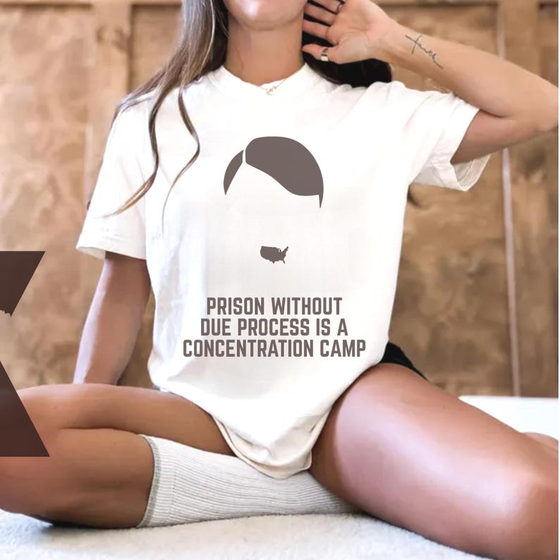 May include: Cream-colored t-shirt featuring a graphic of a person's silhouette and a map of the United States. The text "PRISON WITHOUT DUE PROCESS IS A CONCENTRATION CAMP" is printed below the graphic in a bold font.