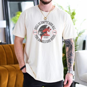 Puede incluir: Camiseta color crema con un dise&ntilde;o gr&aacute;fico circular que muestra un diablo rojo con alas negras. El texto dice "Hell is Empty and All the Devils are Here" y "Shakespeare's The Tempest". La camiseta tiene mangas cortas.