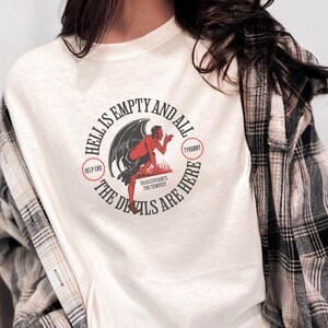 Puede incluir: Camiseta de color crema con un dise&ntilde;o circular que presenta un diablo rojo con alas negras. El texto dice "HELL IS EMPTY AND ALL THE DEVILS ARE HERE" y "TYRANNY". El dise&ntilde;o est&aacute; inspirado en La Tempestad de Shakespeare.