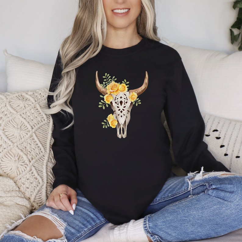 May include: Black sweatshirt with a white floral skull design featuring yellow flowers. The skull has intricate details and is surrounded by green leaves.