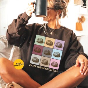 May include: A dark chocolate brown sweatshirt featuring a graphic of nine sourdough loaves in a grid. The text "IN MY SOURDOUGH ERA" is printed below the graphic. A yellow circle with "DARK CHOCOLATE" is in the lower left corner.