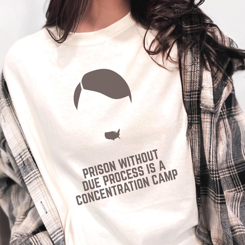 May include: Off-white t-shirt with a graphic of a head silhouette and the outline of the United States. The text "PRISON WITHOUT DUE PROCESS IS A CONCENTRATION CAMP" is printed in dark gray.