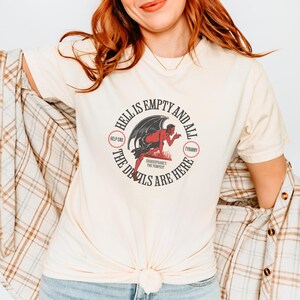 Puede incluir: Camiseta color crema con un dise&ntilde;o gr&aacute;fico circular. El dise&ntilde;o presenta una ilustraci&oacute;n de un diablo rojo y el texto "HELL IS EMPTY AND ALL THE DEVILS ARE HERE." El dise&ntilde;o incluye las frases "HELP END" y "TYRANNY."