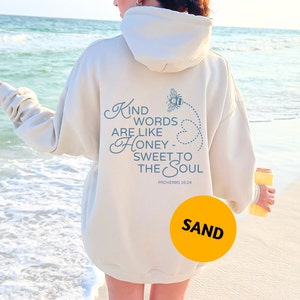 May include: A cream coloured hoodie with a blue bee graphic and the text "Kind words are like honey - sweet to the soul" and "Proverbs 16:24". The hoodie is being worn by a person standing on a sandy beach.